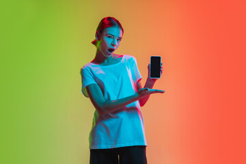 Showing smartphone. Young caucasian girl's portrait on gradient green-orange studio background in neon light. Concept of youth, human emotions, facial expression, sales, ad. Beautiful teen model.