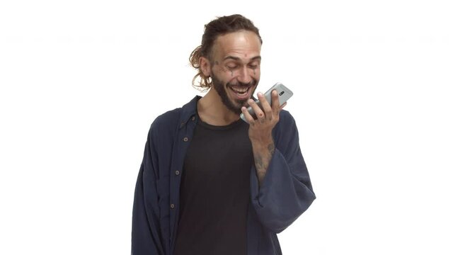 Handsome bearded guy with tattoos on face and body, wearing casual clothing, talking on phone and looking surprised, receive amazing news, smiling happy over white background