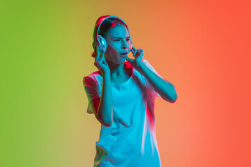 Listening to music with headphones. Young caucasian girl's portrait on gradient green-orange studio background in neon light. Concept of youth, human emotions, facial expression, sales, ad.