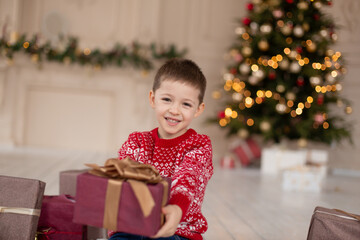 Obraz na płótnie Canvas Portrait of happy smiling little boy in red knitted sweater with Christmas present box near Christmas tree. Happy childhood. Waiting for a Christmas tale