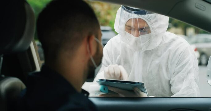 A man is getting temperature measured with infrared thermometer gun by medical staff with PPE suit and list for contact tracking in the case of a coronavirus pandemic at drive thru station.