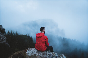 Young bearded hiker wearing red jacket looking at mountain with fog and mist surrounding peaks and...