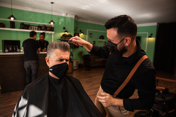 a barber combs an adult male with hand and comb, wears masks pandemic prevention coronavirus, behind two people at the counter talking.