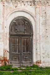 Fototapeta premium Weathered brown wooden gate with cross on top of abandoned brick church near green lawn at countryside on sunny summer day. Traditional architectural style