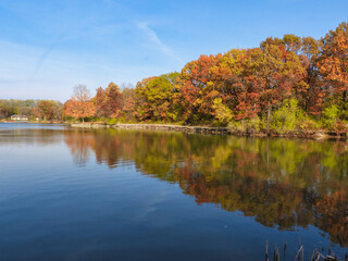 Stunning Fall Scene of Autumn Colored Trees Lining Lake Shore with Boat House to the Side, Bright Blue Sky with Streaking Clouds All Reflected in Calm Water Calm Tranquil October Scene