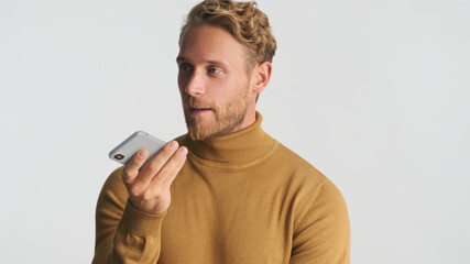 Attractive bearded man wear turtleneck recording voice message for friend on smartphone over white...