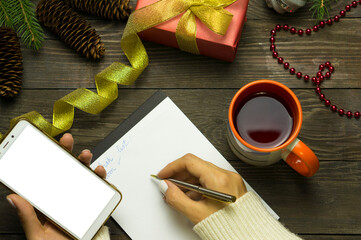 Women's hands in a white sweater make a list on a notebook. On a wooden table there is a cup of tea, a light snack and a notebook with a pen. Wooden table with items symbolizing New Years, Christmas,