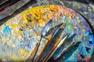 Stock Photo of a painter brushes and palette knife on a wooden palette with mixed and unmixed oil colours on sunlight. Concept art painter