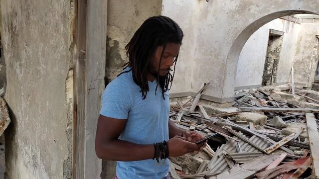 Black African Caribbean dreadlock Rasta man looks at damage and call texts on phone for help in home natural disaster scene - Hurricane, storm, tornado cyclone