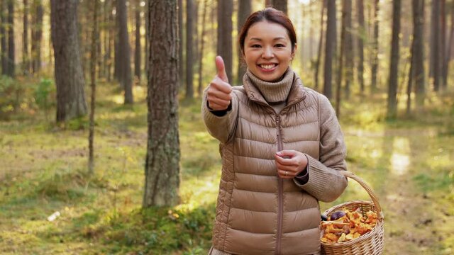 picking season, leisure and people concept - young asian woman with mushrooms in basket showing thumbs up in autumn forest
