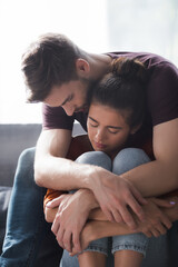 young man embracing and calming beloved woman sitting on sofa with closed eyes