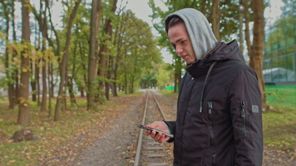 Naklejka na ściany i meble Cropping a view of a millennial hipster, opening and scrolling the phone screen. Young man using a smartphone while walking on the street in the park. A young guy in a hood looks at the phone