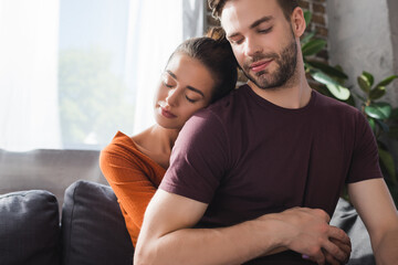 tender woman with closed eyes embracing beloved man from back while sitting on sofa