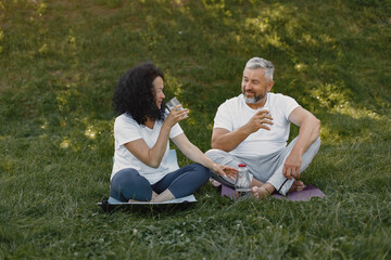 Senior couple is doing yoga outdoors. Stretching in park during sunrise. Brunette in a white t-shirt.