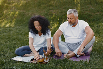 Senior couple is doing yoga outdoors. Stretching in park during sunrise. Brunette in a white t-shirt.