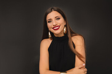 Naklejka premium Smiling lovely girl in black outfit with golden earrings with red lips looking at camera over black backdrop