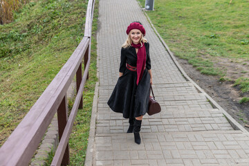 A beautiful woman is walking up the stairs and in a burgundy palette and biret, smiling at the camera in black clothes, in the fall against the background of a pond with blue clouds.