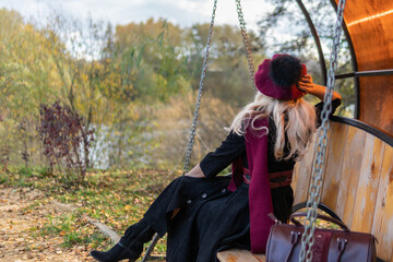 A cheerful lady sits with her back on a garden swing, forehanded in a burgundy coat and in a biret, with beautiful eyes, in autumn against the pond's blue clouds.