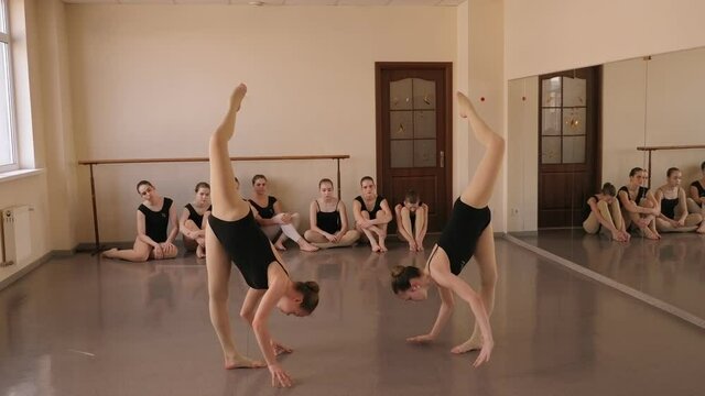 Two flexible young ballerinas are standing in the vertical splits in a ballet class, with ballerinas sitting in the background.