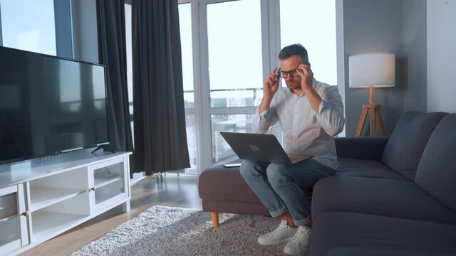 Man with glasses sitting on the couch in a cozy room and using laptop. Remote work concept