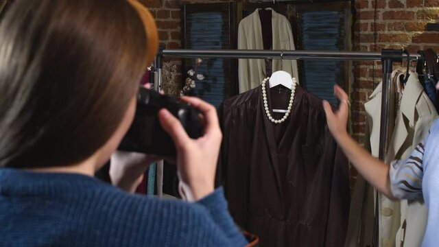 Over the shoulder shot of professional female stylist putting pearl necklace on hanger and holding bag by leather dress while photographer taking pictures for online clothes shop