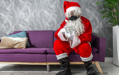 Man disguised as Santa Claus with a mask on Christmas sitting on a couch taking a selfie with his smartphone. Reading the wish list online 