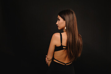 Portrait from back of stylish pretty woman with long hair and golden earrings standing over black background