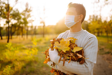 Man in protective medical mask  cleans autumn leaves in the park. Man in gloves collects and piles fallen autumn leaves  in the fall season. Volunteering, cleaning, and ecology concept.