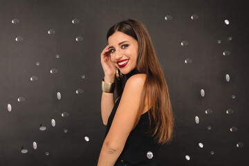 Smiling pretty woman with long dark hair and red lips wearing black outfit posing over black background with confetti
