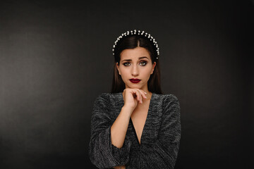 Serious woman with questioning look touching her chin posing over black background with accessorises on the head