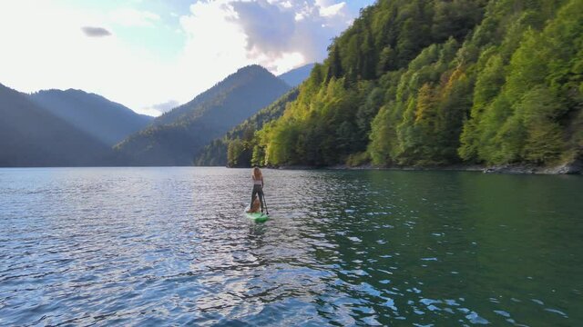 Aerial drone view of pretty fit woman paddle on sup board at mountain lake during sunset with border collie dog. Concept of active tourism. Female silhouette doing water sport during summer holidays
