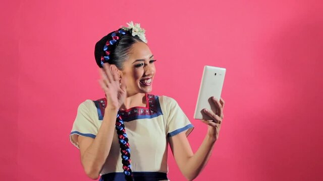 Joven mujer mexicana latina en traje regional folcl&oacute;rico en fondo rosa utilizando tableta para comunicarse redes sociales