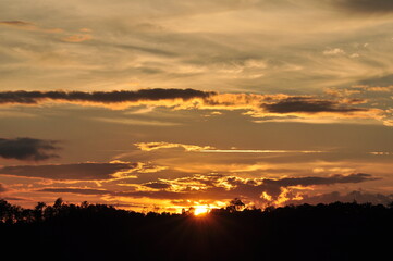 Picture of a sun setting behind a dense forest area followed by mountains.	