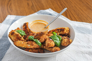 Breaded chicken fingers in a bowl with sauce over wooden table and kitchen towel. Healthy appetiser. Side view. 