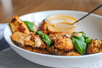 Breaded chicken fingers in a bowl with sauce over wooden table and kitchen towel. Healthy appetiser. Side view. 