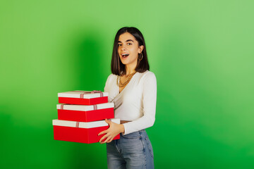 Young joyful woman in casual outfit holding red-white gift boxes on green background. Holidays concept.