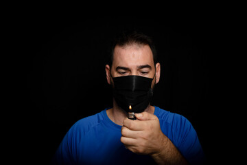 Bearded man dressed in blue t-shirt and with a black mask to protect himself from coronavirus posing against black background
