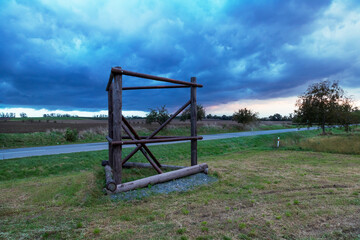 Wooden structure in front of the road in the meadow. In the background is a field and a blue dramatic sky.