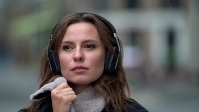 Portrait close-up of a sad beautiful young woman listening to music in black headphones walking through the city on an autumn evening against the background of a road with cars.