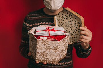 A young man in a medical mask and a red Christmas hat with a gift in his hands, a package of white medical masks and sanitizer. Coronavirus pandemic, quarantine, 2020, 2021. Celebrating the new year