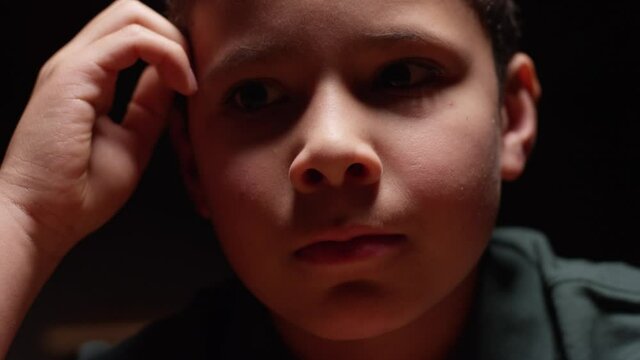 Young hispanic boy sitting at desk in the dark with a sad, lonely expression