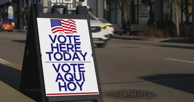 A sign on the sidewalk of a large city shows where to cast your vote. English and Spanish translation.	