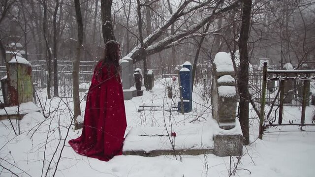 A woman in a red cloak in winter in the cemetery puts flowers on the grave. Headstone in the snow and red carnations