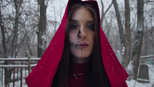 Scary girl in a red hood in winter at the cemetery. Face bodypainting for halloween
