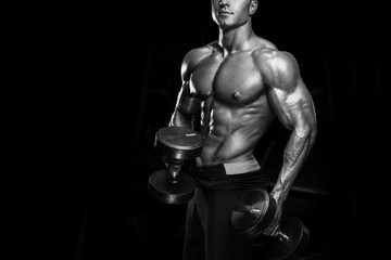 Black and white vertical photo of perfect fit athletic guy workout with dumbbells. Handsome power athletic shirtless man in training pumping up muscles with dumbbells in a gym. Fitness muscular body