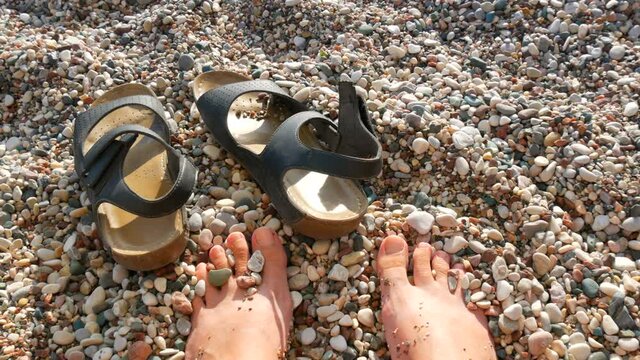 Female toes and leather sandals on the beach of rocky pebble sand on the seashore in summer, vacation concept, POV.