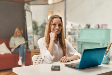 A well-dressed middle age blond hair woman smiling and looking into a camera while sitting at a table in front of a laptop with her colleagues on a background