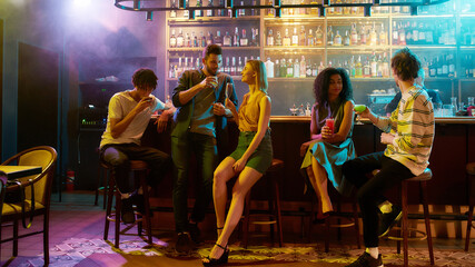 Full length shot of young man leaning on bar counter, drinking cocktail and flirting with woman he just met. Young adults having drinks while chatting at the night club