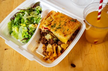 Moussaka with salad in food box, crucible with juice on table.