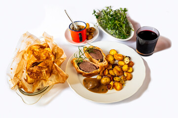 Baked deeer Wellington, portion of meal on plate, cutlery and red wine,pot with sauce and herb.